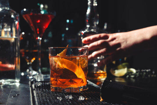 Professional bartender woman serving finished Old Fashioned cocktail with orange zest. Final presentation of classic drink on bar counter mat.