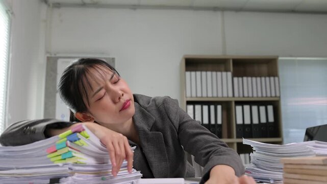 Exhausted young Asian businesswoman feeling sleepy and falling asleep on a pile of paperwork at her office desk from overworking.