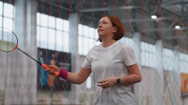 Indoor badminton court practice with woman holding racket near net, pink wristband, white shirt, red hair, calm focused warmup, gentle serves and short rallies, light breathing, community sports hall