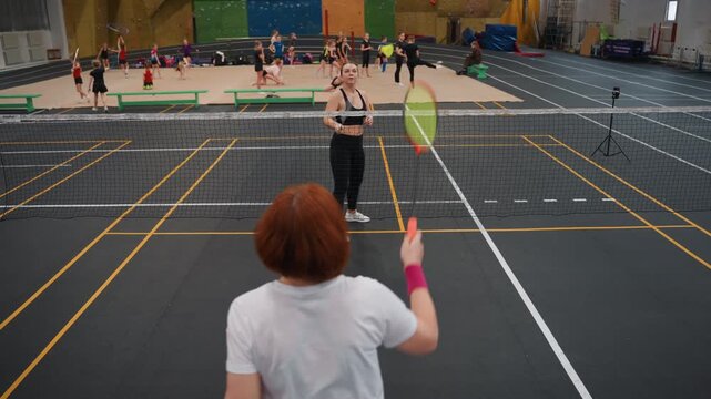 Indoor badminton session. Casual training with focused athlete. Neon wristband worn during badminton practice at gym. Female coach serving softly with trainee near net in sports hall