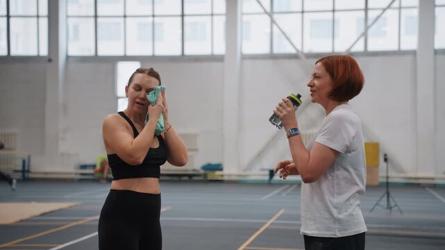 White coach and player postbadminton break wiping faces with towel and sipping water on indoor court, light conversation and strategy notes, supportive mentorship and cooldown moment between drills