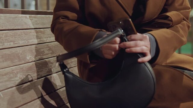 Woman on bench retrieving wallet, anxious urban night scene with leather purse unzipped, fingers searching for card inside zipper pocket, brown coat sleeve and closeup hands revealing tension