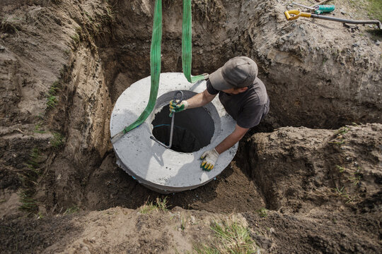 A worker places a concrete septic tank into a hole at a construction site. The worker uses straps for lifting while surrounded by dirt and tools