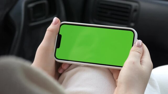 Hands hold phone with green screen inside car. Car interior visible in background. Person wears light-colored clothing. Phone has no brand markings