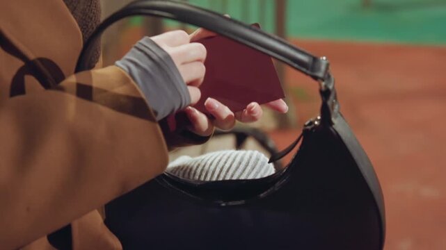 Traveler retrieving burgundy passport from handbag, brown coat and gray fingerless gloves, leather purse with metal clasp, wooden bench at train station, hands flipping passport pages, close detail
