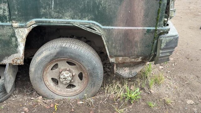 Abandoned dark green minibus with punctured tire, close-up of muddy wheel and spare rim tucked under chassis, heavy rust patches