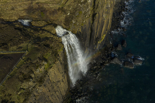 Aerial view of Mealt Falls cascading over the basalt cliffs of Kilt Rock into the dark blue sea with a faint rainbow in the mist Isle of Skye, Scotland, United Kingdom.