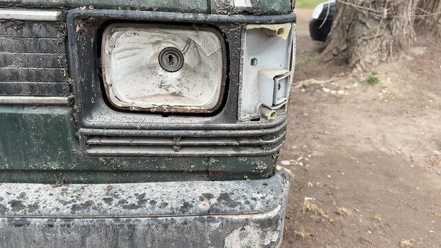 Rusted dark green minibus broken headlight, parked in gravel lot during spring, flaking paint and moss covering corroded body