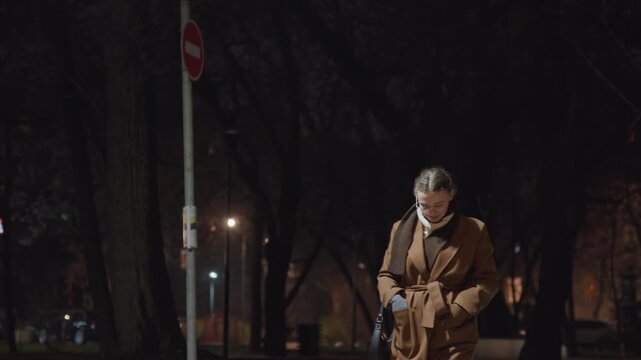 Night park sidewalk with commuter, man in brown coat pauses near lamppost checking phone, visible breath in cold air, silent trees and empty benches, cinematic closeup portrait, contemplative gaze