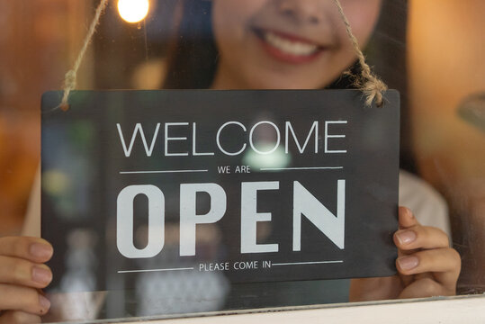 beautiful female business retail owner in medical face mask hanging open wooden sign board at the entrance door of the shop and ready to service customer.