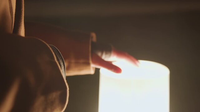Hand touching glowing lamp at night closeup of hand in brown coat reaching over illuminated cylindrical lamp, soft warm light, tactile exploration, urban evening mood, contemplative atmosphere, purse