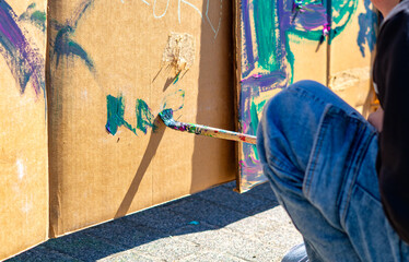 Close-up of a person painting with a brush on a large cardboard sheet outdoors, hand holding a brush with blue paint, person in jeans sitting on pavement, background with colorful paint splashes and g © Adam