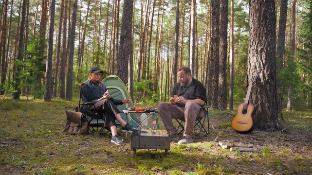 Young couple sitting on folding chairs near a tent, enjoying a barbecue picnic and talking together during a relaxing weekend getaway in a beautiful sunny pine forest