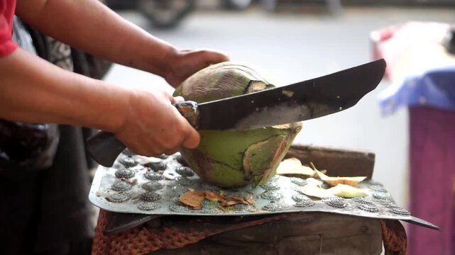 Close-up footage of a woman's hand splitting a fresh young coconut with a wooden-handled machete, a traditional natural health drink of young coconut water in tropical environments.