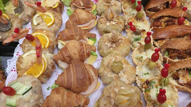 A variety of gourmet snacks on a buffet table, featuring traditional Moroccan chicken or seafood Pastilla (Bastilla) decorated with sliced almonds and physalis. The spread also includes mini croissant