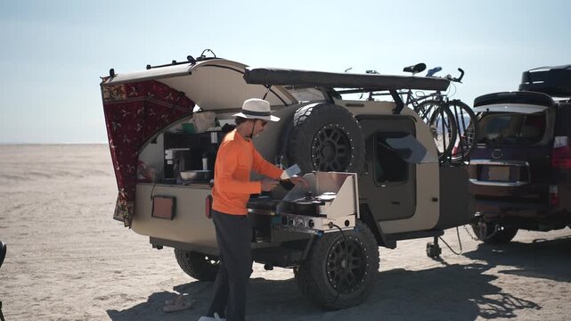 A mature man prepares a meal on a camping stove next to a teardrop trailer at the beach in Baja California, Mexico under blue skies.