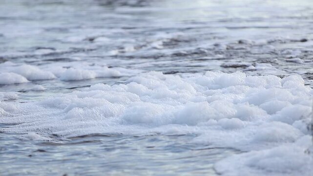 White sea foam floating on calm grey blue water surface with gentle ripples and natural light creating a textured background pattern for environmental or ocean concepts and nature themes