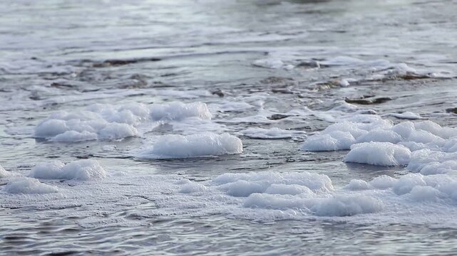 White sea foam floating on calm grey blue water surface with gentle ripples and natural light creating a textured background pattern for environmental or ocean concepts and nature themes