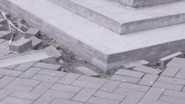 Section of a sidewalk under construction, featuring rough concrete steps leading upwards alongside a partially laid area of grey paving bricks. The scene captures the raw, utilitarian essence of urban