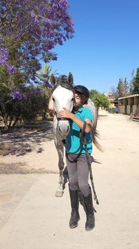 A young boy wearing a black riding helmet and a teal polo shirt stands beside a dapple-gray horse at an equestrian facility, gently stroking the horse's nose. Purple jacaranda tree blossoms frame the 