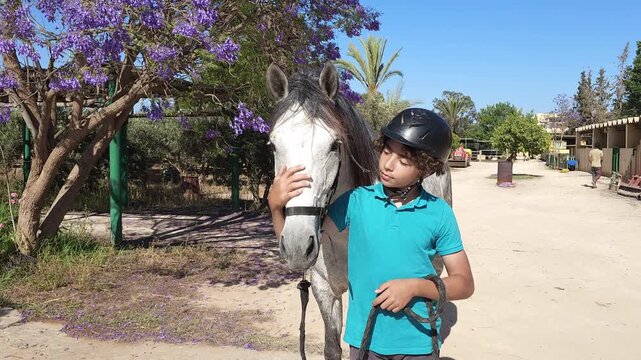 A young boy wearing a black riding helmet and a teal polo shirt stands beside a dapple-gray horse at an equestrian facility, gently stroking the horse's nose. Purple jacaranda tree blossoms frame the 