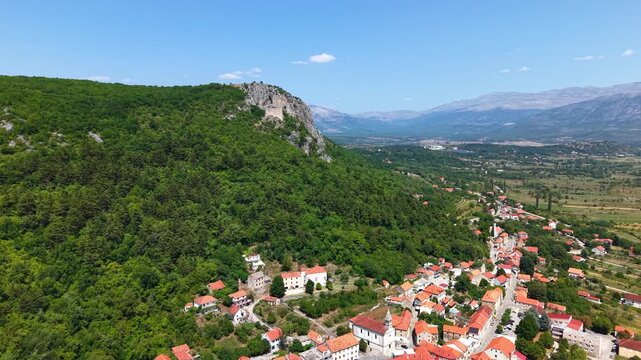 Aerial view of Vrlika town on a rocky hill in the Dalmatian Hinterland, Croatia, featuring red roofed houses and green mountains under a clear blue sky