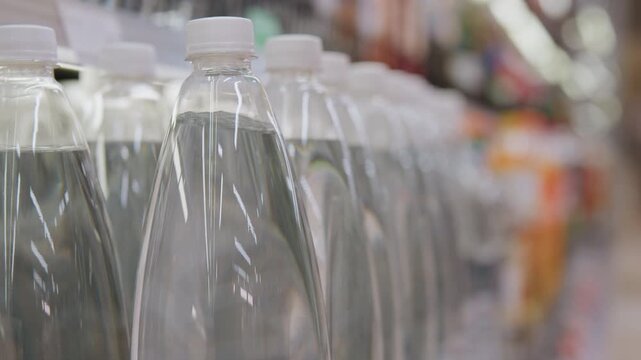Rows of bottled water on shelf, closeup of clear plastic bottles and white caps, supermarket aisle background, shopper hand reaching to pick a bottle, soft lighting and reflective highlights, everyday
