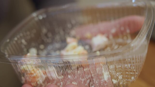 Hands eating leftover salad from plastic container, commuter stirring with white fork in closeup, messy dressing and crumbs visible, midday office lunch captured in disposable clamshell, quick