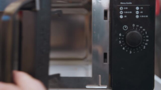 Microwave interior with pizza box placed on tray casual hand slides cardboard box into compact stainless steel oven, closeup of door and control panel, soft kitchen lighting, late night snack scene
