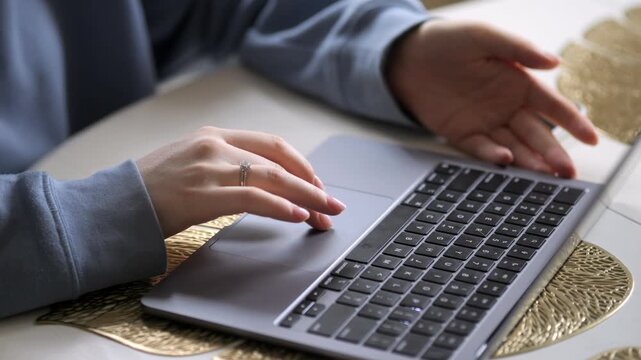 Hands type on laptop keyboard. Person uses touchpad. Golden placemat under device. Light fills room softly