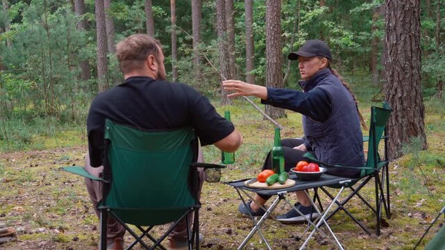 A young couple sits on camping chairs by the fire, drinking beer from green bottles and talking, and behind them in a beautiful pine forest setting there is a tent and a picnic table