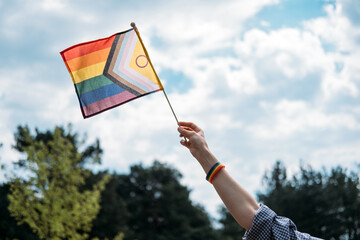 Obraz premium Hand raises progress pride flag against cloudy sky. Human rights advocacy, social justice movement, LGBTQ equality protest.
