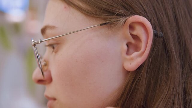 Caucasian teen adjusting glasses profile closeup soft indoor cafe setting, brown hair tucked behind ear, metal frame glasses with chain, freckles visible, fingers brushing hair, focused study vibe,