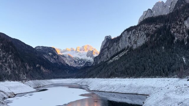 Cinematic sunset over Alpine lake in winter mountains. Winter mountain landscape. Golden hour reflection in calm mountain lake. Alpine peaks illuminated by orange light of setting sun.