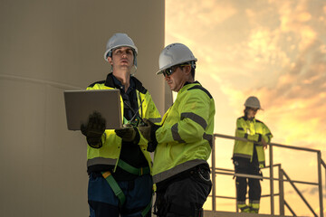 Fototapeta premium Male renewable energy engineers using AI algorithms on laptop to optimize wind turbine performance. Accelerating sustainable power generation at sunset.
