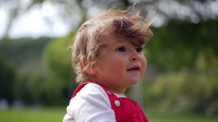 Fototapeta premium Baby in red overalls with wind in hair looking curiously into distance, natural close-up portrait in park conveying innocence, wonder, and early childhood emotion