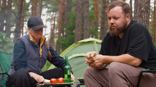 Happy young couple sitting on camping chairs in woods, talking and laughing together, grilling meat on barbecue picnic during their trip to the fresh air and drinking beer