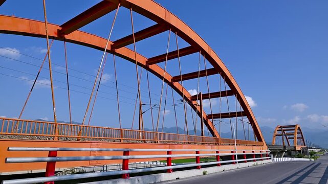 A white high speed train travels across a vibrant red steel arch bridge in Yuli, Hualien, Taiwan, set against a clear blue sky.