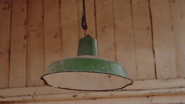 A dusty, chipped green lamp hangs from a ceiling, swaying gently. The light is off, but its presence adds to the rustic ambiance. The wooden planks of the ceiling complete the vintage scene,