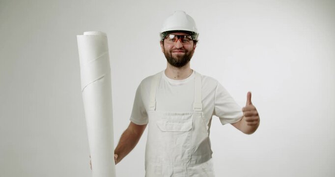 A painter in a white uniform, protective glasses, and helmet holds a roll of wallpaper and gives a thumbs-up against a white background. He smiles at the camera, expressing his satisfaction.