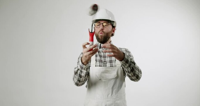 A man wearing a hardhat, glasses, and painter's overalls plays with a paint roller like a microphone. He sings, spins it, and holds it over his eye against a solid white backdrop.