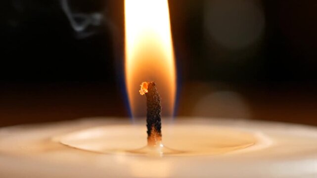 Extreme close-up of a candle wick being ignited by a lighter, showing the orange flame, glowing embers on the charred wick, and a pool of melted wax against a dark background.