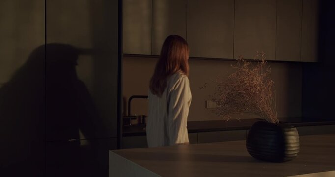 Woman walks into a modern, minimalist kitchen in the morning. She is wearing pajamas and is about to start her day. Warm sunlight illuminates the room, creating a cozy and inviting atmosphere.