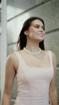 A stylish woman in a pink dress opens a green door, steps out, and smiles while walking down the street. Her pearl necklace complements her elegant look as she confidently navigates the urban city