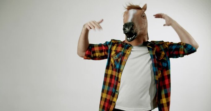 A man wearing a horse mask dances against a white background. He is wearing a colorful plaid shirt and a white t-shirt. He moves his body and arms to the rhythm of music which