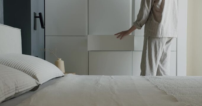 Woman in pajamas opens a drawer in a modern bedroom. The room features minimalist design with white furniture and dark walls. The bed is made with white linens.