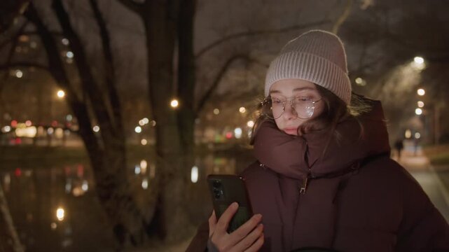 Night park bench white woman calling phone, closeup portrait with beanie and glasses, muffled breath in cold air, soft bokeh lights over river, hand gestures and thoughtful expression, candid urban