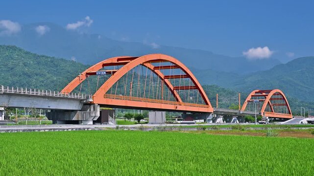 A white passenger train travels across a vibrant red arch bridge surrounded by lush green rice fields and mountains in Yuli, Hualien, Taiwan.