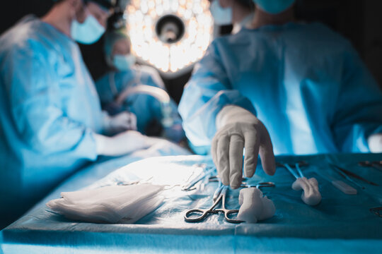 Close up of assistant hand reaching for surgical instruments during operation, blurred background with surgeons performing procedure, sterile environment, medical teamwork and healthcare concept