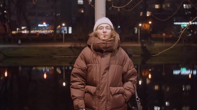 Lakeside night white woman smoking contemplative, wearing puffer jacket and beanie with glasses, closeup exhale over water reflections and city lights, intimate portrait of latenight thinker